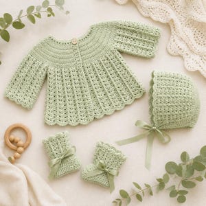 Puede incluir: Conjunto de bebé de ganchillo verde claro, que incluye una chaqueta, un gorro y patucos, todos con un delicado diseño calado. El conjunto presenta un borde festoneado y está atado con cintas de raso. También hay un mordedor de madera.