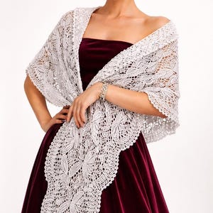 May include: A white, open-weave shawl with a floral pattern. The shawl is draped over a burgundy velvet dress. The intricate lace design is visible, creating a delicate and elegant look. The shawl is a beautiful accessory.
