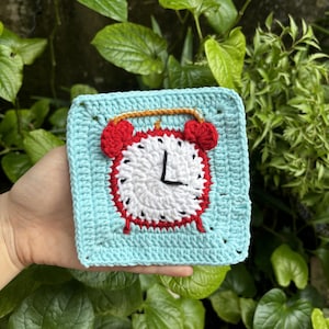May include: A crocheted square with a red and white alarm clock design. The clock face has black numbers and a black hand pointing to the 12. The square is a light blue color.