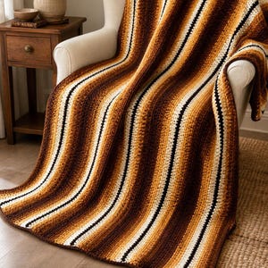 May include: A crocheted blanket with a striped pattern in shades of brown, tan, and cream. The blanket is draped over a white armchair, showcasing its texture and design. The stripes run vertically, creating a cozy and inviting aesthetic.