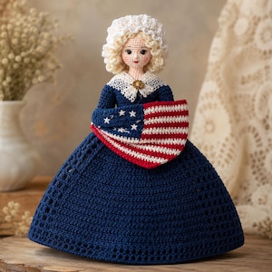 May include: A handcrafted crochet doll with blonde curly hair wearing a white bonnet, a dark blue dress with a white lace collar, and a small brooch. The doll holds a miniature American flag featuring red and white stripes and a blue field with white stars.