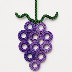Pode incluir: Um ornamento decorativo suspenso em forma de cacho de uvas, feito com anéis de crochê roxos dispostos em um cacho. A peça apresenta um caule de crochê verde e duas folhas verdes encaracoladas no topo, suspensa por um cordão de corda verde.