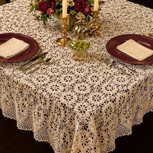 Peut inclure: Une nappe en crochet de couleur crème avec un motif floral répétitif et des bords festonnés. La table est dressée avec deux assiettes rouge foncé, des serviettes beiges, des couverts en argent, deux bougeoirs en laiton et un bol en verre.