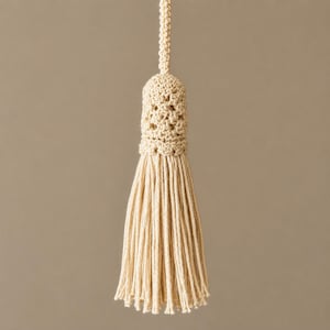 May include: Cream-colored, handmade tassel with a crocheted top and long, dangling strands. The tassel is suspended against a neutral background, showcasing its texture and design. This decorative element is suitable for various craft projects.