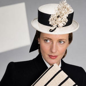 May include: A white hat with a black band and floral embellishments. The hat has a flat brim and a cylindrical crown. A black fabric extends from the hat. The woman is wearing a black blazer and holding a cream-colored clutch with black stripes.