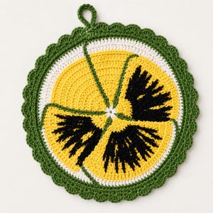Pode incluir: Um descanso de panela circular em crochê com um design de flor amor-perfeito. O centro é branco, rodeado por pétalas amarelas com padrões pretos radiantes. A borda externa é finalizada com um acabamento verde festonado e uma pequena alça.