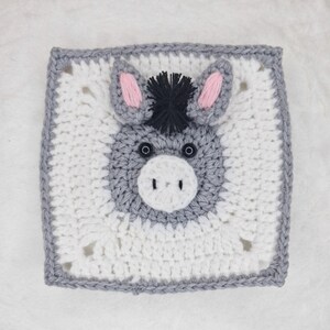 May include: A square, crocheted donkey face with a white background and gray border. The donkey has gray fur, pink ears, black eyes, and a black mane. The donkey's face is centered on the square.