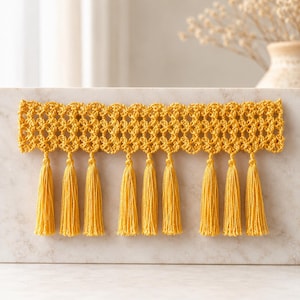 Pode incluir: Uma guarnição de crochê amarelo-mostarda com um intrincado padrão de treliça aberta ao longo da borda superior e uma série de nove borlas pendentes correspondentes fixadas na base, criando uma borda têxtil decorativa de tom dourado.