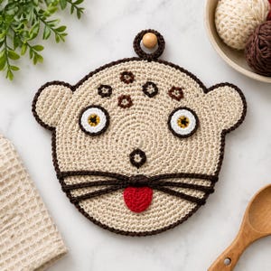 May include: A beige, crocheted pot holder in the shape of a cat's face. The cat has brown whiskers, a red tongue, and brown accents around the eyes and head. A wooden bead is attached for hanging. The pot holder is round and has a circular pattern.