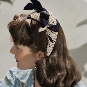 May include: A vintage-style head covering made of cream-colored crocheted material, adorned with black satin bows. The head covering is worn by a person with brown hair and a blue floral patterned top. The person is wearing gold hoop earrings.