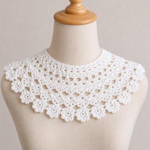 May include: A white crochet collar featuring an intricate openwork pattern with a scalloped edge composed of small floral motifs. The accessory is designed to sit around the neck and shoulders, showcasing detailed needlework and a delicate texture.