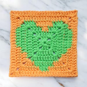 May include: A crocheted square with a green heart design in the center. The square is surrounded by a border of orange crochet.