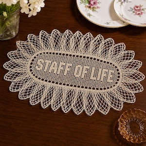May include: An oval-shaped beige crochet doily featuring an intricate openwork mesh border and a central rectangular panel with the text "STAFF OF LIFE" woven into the fabric using a dense, solid stitch pattern against the delicate lace background.