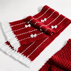 Pode incluir: Um cachecol de crochê vermelho escuro com linhas horizontais costuradas em branco e pequenos laços de fio branco fixados em intervalos. As extremidades do cachecol são finalizadas com longas franjas brancas, conferindo textura ao tecido.