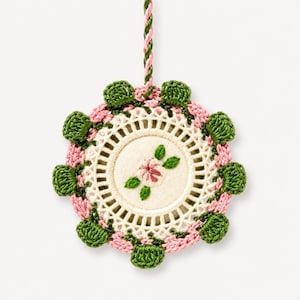 Pode incluir: Um ornamento circular suspenso com um disco central de tecido creme apresentando uma pequena flor rosa bordada e folhas verdes. O disco é rodeado por uma borda de crochê creme, detalhes em fio rosa e uma borda festonada de crochê verde.