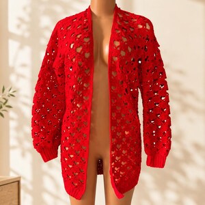 Puede incluir: Un cárdigan rojo brillante abierto por delante que presenta un intrincado patrón de ganchillo con recortes repetidos en forma de corazón en todo el cuerpo y las mangas, acabado con puños acanalados y un dobladillo recto en la parte inferior.