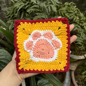 May include: A crocheted square with a pink paw print in the center. The paw print is surrounded by a white border and the square is bordered in red and yellow.