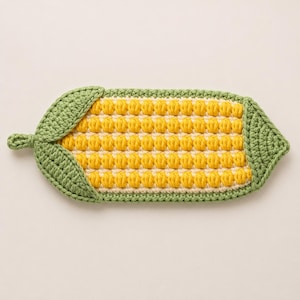 May include: A crochet corn cob shaped coaster or trivet featuring a textured yellow kernel pattern made with bobble stitches, framed by light green husks on both ends, and finished with a small loop on the left side for hanging or storage.