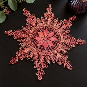 May include: A circular lace doily featuring a vibrant coral and orange gradient color scheme. The design showcases a central floral motif with eight petals surrounded by an intricate web of delicate, star-shaped, and pointed lace patterns.