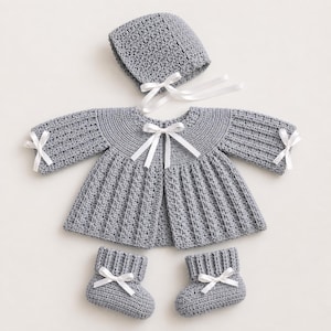 May include: A gray crochet baby set including a jacket, bonnet, and booties. The jacket and bonnet have white ribbon ties, and the booties also feature white ribbon accents. The set is made of a textured, soft yarn.
