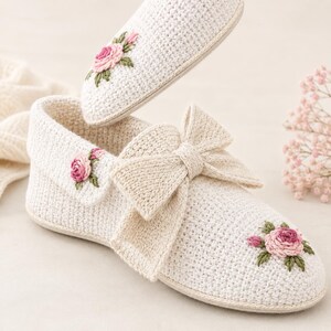 May include: A pair of off-white crochet slippers featuring a large bow on the front and embroidered pink rose motifs with green leaves. The slippers have a textured knit construction and a rounded toe design, showcasing delicate floral detailing.