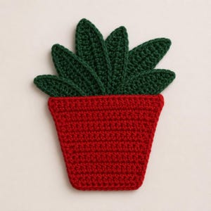 May include: A crochet coaster shaped like a potted plant. The base is a trapezoidal red pot with a textured stitch pattern. Emerging from the top are several pointed, dark green leaves arranged in a fan shape, also crafted in crochet.