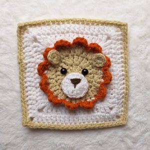 Puede incluir: Un rostro de león tejido a ganchillo, con melena y cara beige, melena naranja, ojos negros y hocico blanco, centrado en un cuadrado blanco con borde beige. El tejido tiene un aspecto texturizado.