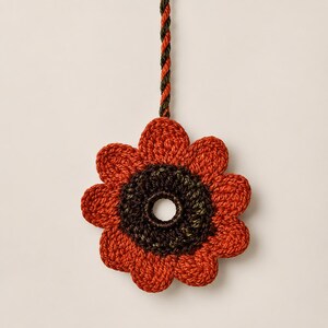 Pode incluir: Um ornamento de flor em crochê com dez pétalas arredondadas em fio laranja queimado vibrante. O centro é um anel circular texturizado castanho escuro com uma abertura oca. Um cordão torcido em castanho e laranja estende-se para cima.