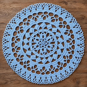 May include: A round, light blue crochet doily featuring a complex, symmetrical pattern of interconnected loops, solid stitches, and openwork sections. The design radiates from a central floral motif outward to a scalloped, decorative edge.