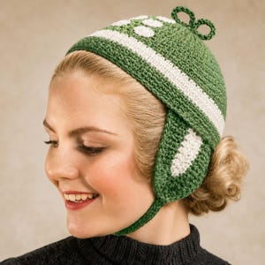 May include: A green crochet hat featuring a white horizontal stripe, white circular accents, and a small decorative loop at the top. The hat includes ear flaps that extend downward and are secured with a thin crochet strap under the chin.