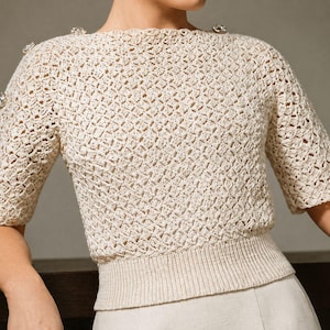 May include: Cream-colored crochet top with a boat neck and short sleeves. The top features decorative buttons on the shoulders and sleeves. The crochet pattern has a diamond-like design. The bottom hem is ribbed.