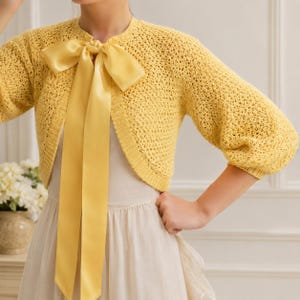 May include: A bright yellow cropped cardigan featuring an open-knit texture, three-quarter length sleeves, and a ribbed neckline. The garment is secured at the front with a long, wide satin ribbon tied into a large bow at the center of the chest.