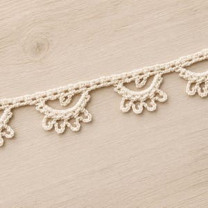 May include: A cream-coloured crochet lace trim featuring a repeating scalloped edge pattern. The trim consists of a straight woven header band with decorative semi-circular motifs extending downward, each finished with small, delicate loops.
