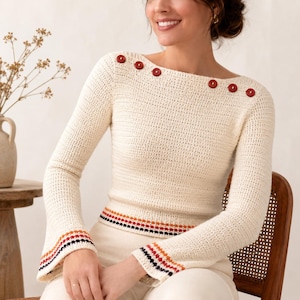 May include: Cream-colored crochet sweater with long sleeves and a boat neck. The sweater features six red buttons along the neckline and a striped design at the waist and cuffs. The sleeves have a flared design.