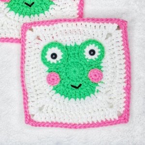 May include: A white crocheted square with a pink border features a green frog face with black eyes and pink cheeks.