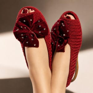 Puede incluir: Un par de zapatillas rojas de ganchillo con diseño de punta abierta. Cada zapatilla presenta un gran lazo de terciopelo rojo oscuro adornado con brillantes pedrería. Las zapatillas tienen un aspecto texturizado y un diseño cómodo y elegante.