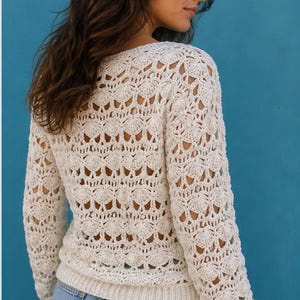 May include: Cream-colored crochet sweater with a detailed openwork pattern. The long-sleeved top features a round neckline and ribbed cuffs and hem. The sweater is paired with light blue denim jeans. The background is a solid teal color.