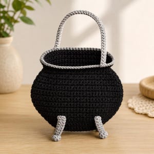 Puede incluir: Una cesta de ganchillo negra con forma de caldero, con un borde gris, un asa arqueada gris y tres patas de ganchillo grises. La cesta presenta un cuerpo redondeado y una abertura ancha, construida con un patrón de puntos consistente.