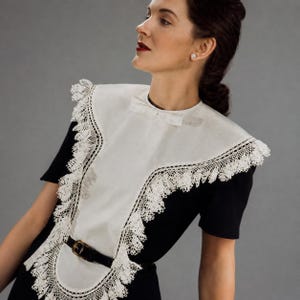 Peut inclure: Un col amovible blanc orné d'une bordure en dentelle complexe et d'un petit nœud à l'encolure. Le col est porté sur un vêtement noir à manches courtes, resserré à la taille par une fine ceinture noire dotée d'une boucle dorée.