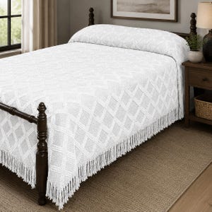 May include: A white bedspread featuring a textured diamond pattern across the surface and long fringe detailing along the edges. The textile is draped over a dark wooden bed frame, covering the mattress and extending down to the floor on the sides.