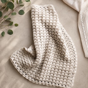 May include: A cream-colored, crocheted blanket with a textured, honeycomb-like pattern. The blanket is draped, showcasing its soft, flowing form. A portion of a matching sweater is visible in the upper right corner, suggesting a coordinated set. The blanket is approximately 60 inches long.