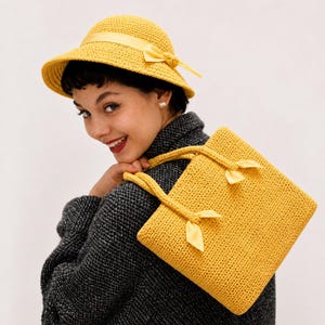Puede incluir: Un sombrero amarillo tejido a ganchillo con una cinta y un bolso tote amarillo tejido a ganchillo a juego con detalles de cinta. La mujer lleva un abrigo gris oscuro. El sombrero y el bolso son de un amarillo brillante y soleado.