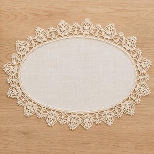 May include: An oval-shaped off-white doily featuring a smooth fabric center surrounded by an intricate, wide crocheted lace border with a repeating pointed pattern. The delicate textile piece displays a consistent cream color throughout the design.