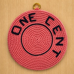 May include: A circular, coral-colored crocheted wall hanging featuring the black embroidered text ONE CENT arranged in a circular pattern. The piece includes a small, gold-toned metal ring at the top center for hanging purposes.