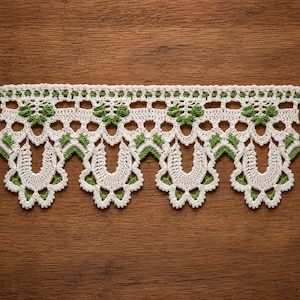 May include: A decorative crochet lace trim featuring a repeating pattern of off-white yarn and small green accents. The design includes a straight top edge with a series of openwork stitches and a scalloped bottom edge with U-shaped motifs.