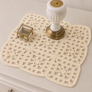 May include: A square, off-white crochet doily featuring a repeating floral motif within a grid pattern. The doily has a scalloped edge and is constructed from interconnected yarn squares, creating an openwork design with a textured, knotted appearance.