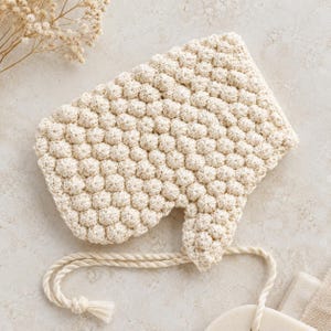 May include: A cream-colored crocheted bonnet featuring a textured bobble stitch pattern throughout the fabric. The headwear includes a rounded shape with a small brim section and two long, twisted rope-style ties attached at the base for fastening.