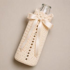 May include: A clear glass bottle encased in a cream-colored crocheted sleeve featuring a vertical open-stitch pattern. The sleeve is secured at the neck with a smooth, matching cream-colored satin ribbon tied into a bow with two long hanging ends.