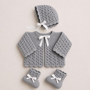 May include: A gray crochet baby set, including a jacket, bonnet, and booties, each adorned with white satin bows. The set features a scalloped edge design and is made of soft, textured yarn. A complete outfit for a newborn.