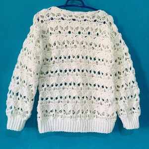 May include: A white, open-weave crochet sweater with long sleeves and a ribbed hem and cuffs. The sweater is hanging on a blue hanger against a blue background. The crochet pattern features a repeating shell design.